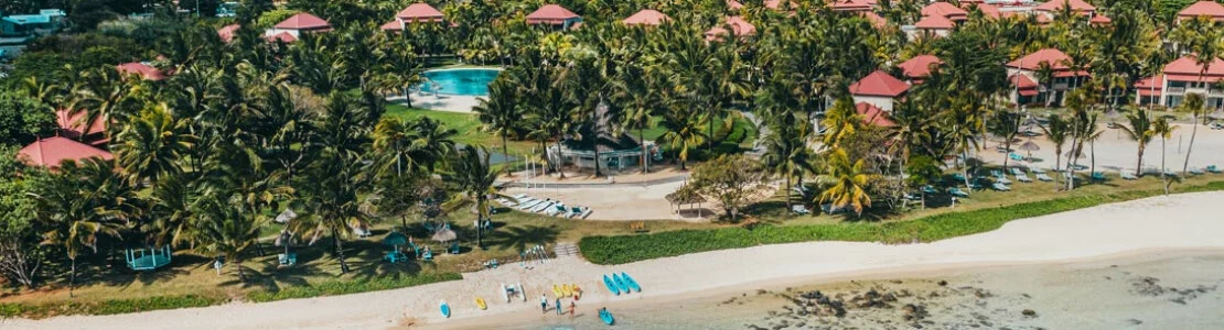 Palm-lined resort grounds with a turquoise pool, red-roofed buildings, and a sandy beach.