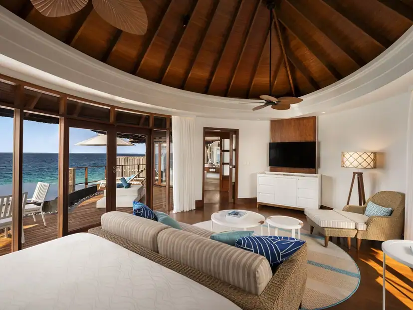 Constance Halaveli: A bright villa living area with ocean-facing doors opening onto a furnished wooden deck.