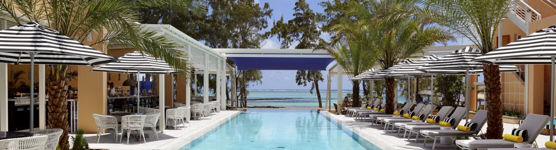 A palm-lined outdoor pool is surrounded by sun loungers and striped parasols, with the ocean visible beyond.